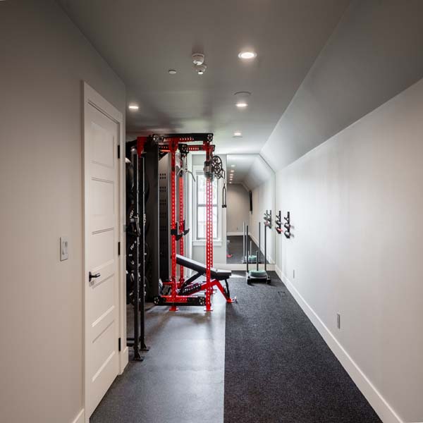 Riddle_Construction_Water_Hazard_Weight_Room Narrow home gym along a hallway featuring a red power rack, weight plates, a bench, and a mirror at the far end; coat hooks line the right wall.